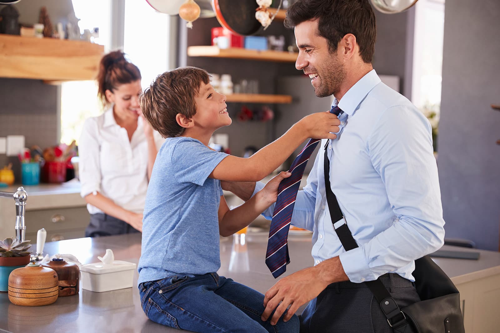 Young boy helping father tie necktie in modern kitchen before work or school.