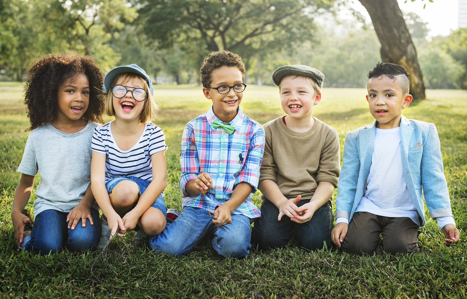 Five happy children sitting together on grass in a park, smiling at camera.