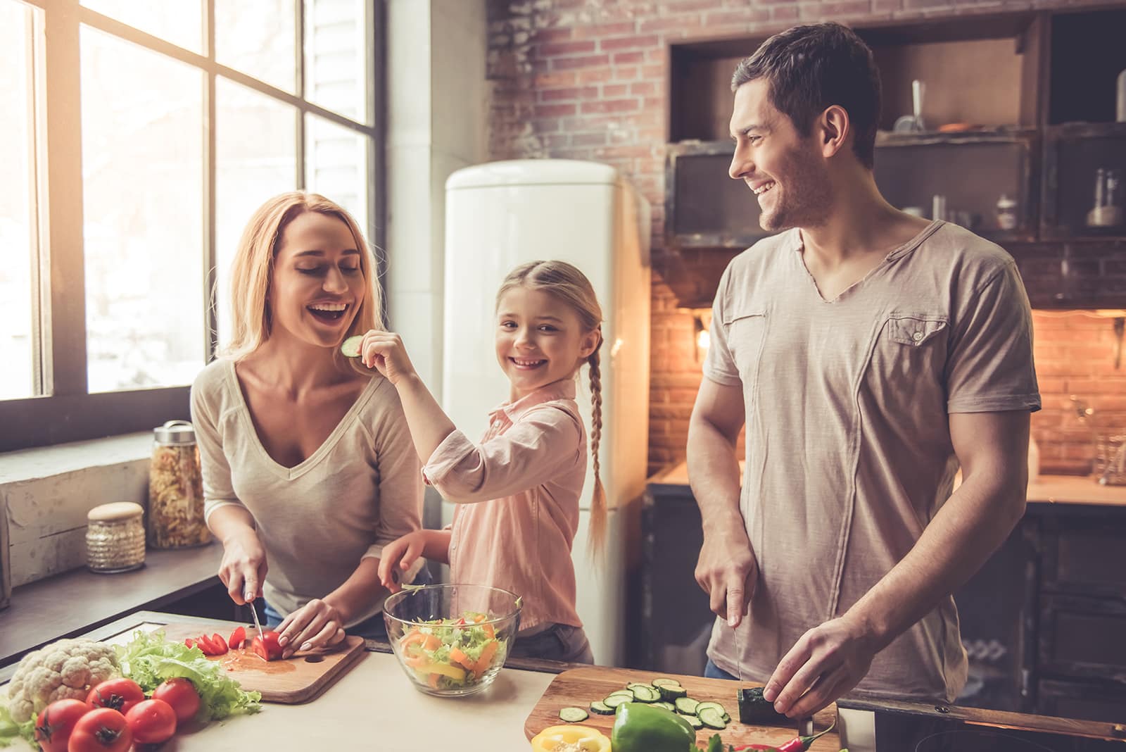 Family of three cooking together in bright kitchen with fresh vegetables on counter.
