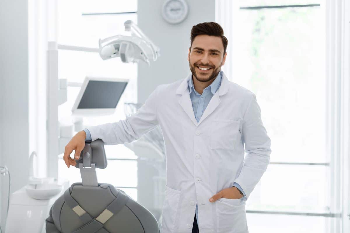 Smiling dentist in white coat standing in modern dental office beside equipment.