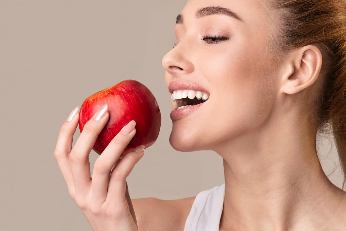 Woman smiling while holding red apple near mouth against neutral beige background.
