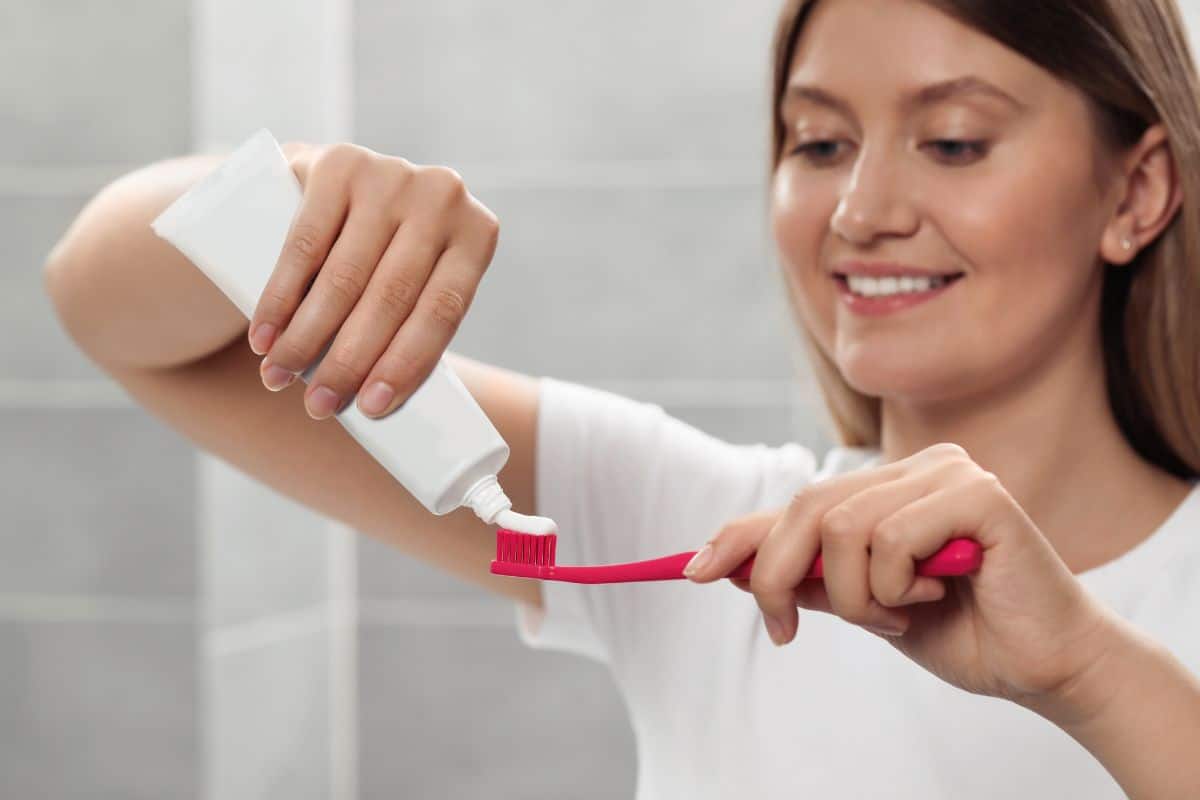 Does-Whitening-Toothpaste-Work Woman applying toothpaste to pink toothbrush in bathroom setting.
